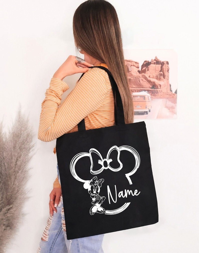May include: Black tote bag with a white outline of Minnie Mouse's head and bow. The bag has the word "Name" written below the image.