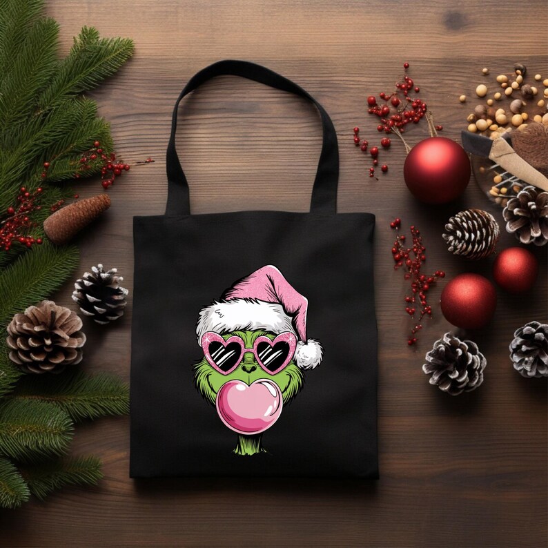 May include: Black tote bag featuring a green cartoon character wearing a pink Santa hat and heart-shaped sunglasses, blowing a pink bubble. The bag has black handles and is set against a wooden background with Christmas decorations.