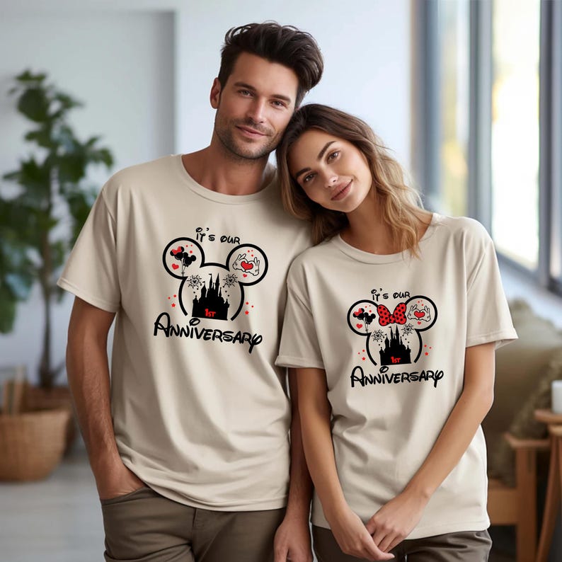 May include: Two beige t-shirts featuring a black Mickey Mouse head design. The design includes a castle, hearts, and the text "It's our 1st Anniversary". One shirt has a red bow, the other has a red heart.