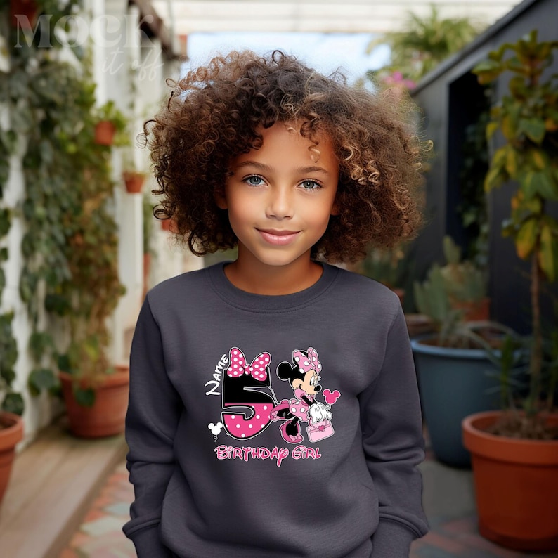 May include: A dark gray sweatshirt with a pink and white Minnie Mouse design. The design features Minnie Mouse holding a pink gift bag and a pink heart balloon. The number 5 is in the design and the text "Name Birthday Girl" is below the design.