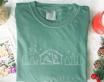 May include: A sage green Comfort Colors t-shirt featuring a white line drawing of a nativity scene. The design includes an angel, stable, and figures. The shirt has a crew neck and a tag that reads "Comfort Colors".
