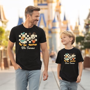 May include: Black t-shirts with a cartoon graphic of Donald Duck and Mickey Mouse on a checkered background. The shirts have the text "Big Trouble" and "Little Trouble" in a playful font. The shirts are worn by a father and son.