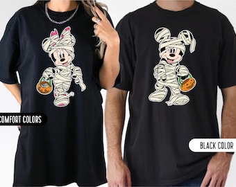 May include: Two black t-shirts featuring graphic prints of cartoon characters wrapped in white bandages like mummies. One character wears a pink bow and holds an orange jack-o'-lantern bucket, while the other holds a similar pumpkin bucket.