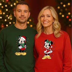 K&ouml;nnte beinhalten: Zwei Personen tragen passende Weihnachtspullover. Einer ist dunkelgr&uuml;n, der andere rot. Jeder Pullover zeigt eine Cartoonfigur mit Weihnachtsm&uuml;tze und Lichterketten. Der Hintergrund ist ein Weihnachtsbaum.