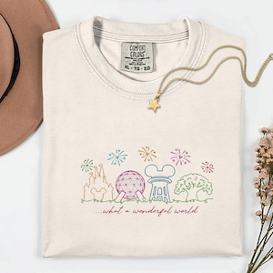 May include: Cream-colored Comfort Colors t-shirt with a colorful line drawing of a castle, Spaceship Earth, and other landmarks. The text "...what a wonderful world" is below the drawing. A gold star pendant necklace is on the shirt.