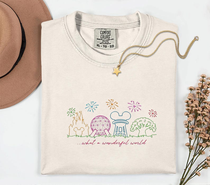May include: Cream-colored Comfort Colors t-shirt with a colorful line drawing of a castle, Spaceship Earth, and other landmarks. The text "...what a wonderful world" is below the drawing. A gold star pendant necklace is on the shirt.