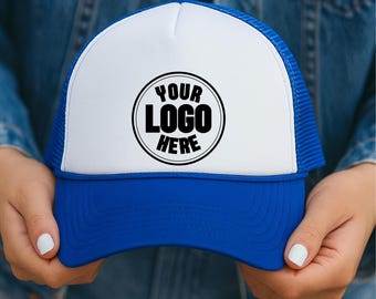 May include: A blue and white trucker hat with the text "YOUR LOGO HERE" in a black circle. The hat has a blue brim and mesh back, with a white front panel. The hat is held up by a person.