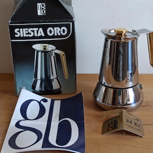 May include: A silver and gold Siesta Oro coffee maker with a gold lid and handle. The coffee maker is sitting on a wooden surface with a white paper with the letters "gb" printed on it. The coffee maker is in front of a black box with the words "Siesta Oro" printed on it.