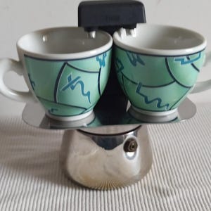 Puede incluir: Una cafetera italiana de acero inoxidable con dos tazas a juego. Las tazas son blancas con un diseño abstracto en verde menta y azul. La cafetera tiene un asa negra y una base plateada.