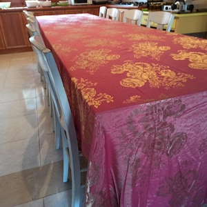 May include: A long, rectangular, red tablecloth with a gold floral pattern. The tablecloth is draped over a table with several white chairs.