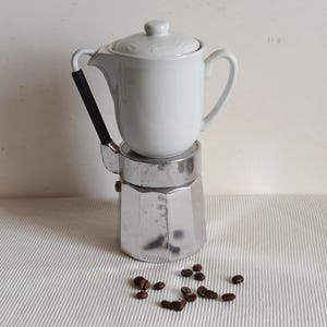 Puede incluir: Una cafetera de cerámica blanca con una base plateada y un asa negra. La cafetera está sobre una superficie blanca con granos de café esparcidos alrededor.