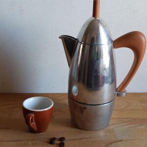 Puede incluir: Una cafetera italiana de acero inoxidable con asa y parte superior marrones, junto a una pequeña taza de espresso marrón. Granos de café esparcidos cerca de la taza. La cafetera mide aproximadamente 23 cm de alto.