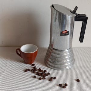 May include: Silver coffee pot with a black handle and a red and white label that says "Vev Vigano". A brown ceramic coffee cup and coffee beans are on a white surface.