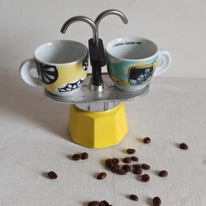 May include: A yellow stovetop espresso maker with two small white coffee cups. One cup has a yellow and black design, the other has a picture and the text "I Labrini Espresso 1996". Coffee beans are scattered around the base.