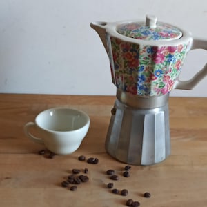 Könnte beinhalten: Ein Espressokocher mit Blumenmuster und weißem Griff und Deckel, neben einer kleinen weißen Tasse. Kaffeebohnen sind auf der Holzoberfläche verstreut. Der Espressokocher hat einen silbernen Sockel.