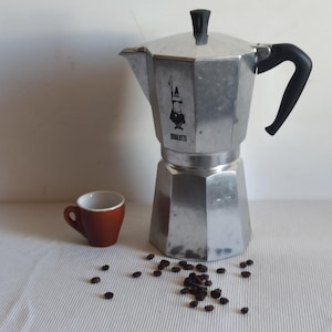 May include: A silver Bialetti stovetop espresso maker with a black handle and a small white cup with brown coffee beans scattered around it.