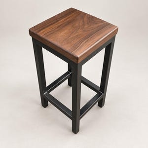 May include: A square bar stool featuring a dark brown wooden seat with a visible grain pattern. The base is constructed from four straight black metal legs connected by a square support frame at the lower section, providing a stable structure.