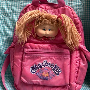 May include: A pink Cabbage Patch Kids backpack with a doll face and yarn hair attached. The backpack has a front pocket with a zipper and the Cabbage Patch Kids logo. The backpack has a top handle and adjustable straps.