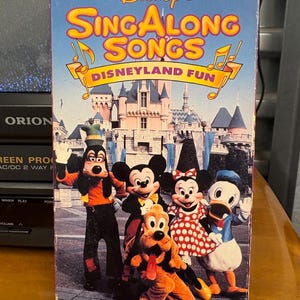 May include: A vintage Disney's Sing Along Songs VHS tape, Volume Seven, featuring Disneyland Fun. The cover displays Mickey Mouse, Minnie Mouse, Goofy, Donald Duck, and Pluto in front of a castle. The title is in yellow and orange.