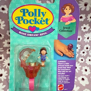 May include: A Polly Pocket Rose Dream Ring toy set in its original packaging. The set includes a ring with a clear, hinged top, a small doll, and the text "Jewel Collection". The packaging is teal and purple.