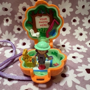 May include: An open, orange Polly Pocket playset with a miniature figure wearing a green hat and gray outfit. The playset features a tree, a swing, and the text "YOUR PHOTO HERE". A purple lanyard is attached.