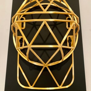 May include: A gold-colored wire frame hat rack mounted on a black background. The hat rack is shaped like a baseball cap.