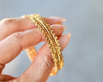 May include: A gold chain bracelet with a woven design. The bracelet is made of small, square-shaped links that are interlocked to create a textured pattern.