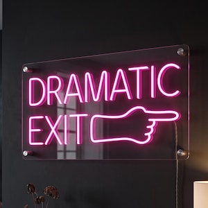 May include: A rectangular clear acrylic wall sign featuring bright pink neon-style lettering that reads DRAMATIC EXIT with a pointing hand icon. The sign is mounted with four silver hardware standoffs and includes a visible power cord hanging down.