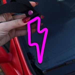 Pode incluir: Um chaveiro em forma de raio rosa com uma correia preta. O chaveiro está pendurado no espelho retrovisor de um carro.