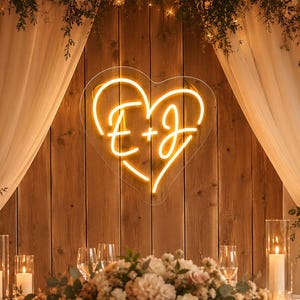 Può includere: Un'insegna al neon a forma di cuore con le lettere gialle illuminate E e J separate da un segno più. L'insegna è montata su una parete di assi di legno verticali, sopra un tavolo decorato con composizioni floreali e candele.