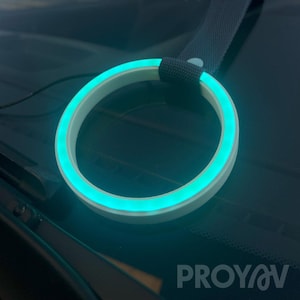 Può includere: Un accessorio per auto circolare e luminoso con un anello esterno bianco e una luce interna blu brillante. L'accessorio è attaccato a una cinghia grigio scuro. La parola "PROYAV" è stampata in bianco nell'angolo in basso a destra dell'immagine.