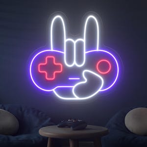 Puede incluir: Un letrero de neón con forma de mando de videojuego con un gesto de la mano, con un signo más y un círculo rojos. El letrero está delineado en blanco y morado, con un fondo oscuro. Una pequeña mesa de madera y pufs están en primer plano.