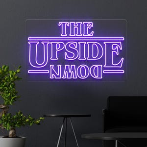Puede incluir: Un letrero de neón rectangular con las palabras "THE UPSIDE DOWN" en púrpura. El letrero está montado en una pared gris oscuro, con una pequeña planta y una mesa negra en primer plano. Es una referencia a una serie de ciencia ficción.
