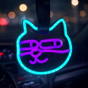 May include: A cat-shaped car accessory featuring a glowing cyan neon outline and a purple neon graphic of a cat face with squinted eyes and a nose. The item is suspended by a black fabric strap from a rearview mirror in a dark vehicle interior.