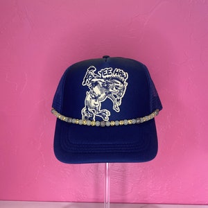 May include: A navy blue trucker hat with a white embroidered design of a cowboy riding a bucking bronco. The hat has a white and gold beaded trim around the brim. The text "Yee Haw" is embroidered on the hat.