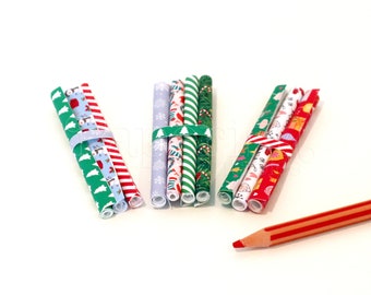 May include: Three bundles of rolled decorative paper with festive patterns, including snowflakes, candy canes, and Christmas trees. Each bundle is secured with a matching elastic band. A red and orange striped pencil rests on the white surface.