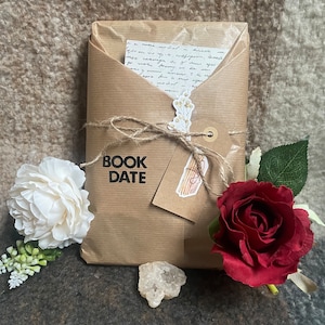 Op de afbeelding: Een in bruin papier verpakt pakket, vastgebonden met touw, met een label met de tekst "BOOK DATE". Een witte bloem, een rode roos en een klein kristal zijn eromheen gerangschikt. Een handgeschreven notitie is zichtbaar.