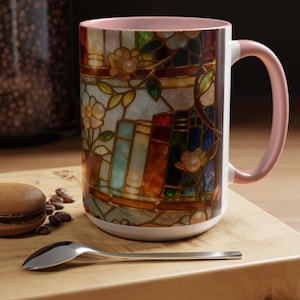 May include: A white ceramic mug with a stained-glass design featuring books, flowers, and a pink handle. The mug is decorated with warm colors like orange, brown, and blue. A macaron, coffee beans, and a spoon are on a wooden surface.
