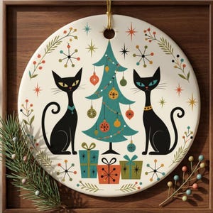 May include: A round ceramic ornament featuring two black cats flanking a teal Christmas tree decorated with colorful ornaments. Below the tree are three gift boxes in teal, orange, and green. The background is cream with starbursts and sprigs.