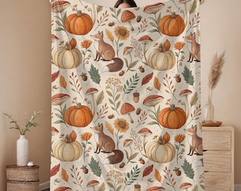 May include: A cream-colored throw blanket featuring a repeating pattern of orange pumpkins, brown foxes, various mushrooms, sunflowers, acorns, and autumn leaves. The design is distributed across the fabric in a balanced, decorative arrangement.