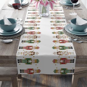 May include: White table runner featuring a repeating pattern of festive nutcracker figures in red, green, and gold. The design includes stars and is set against a white background. Perfect for holiday decor.