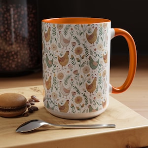 May include: White ceramic mug with an orange interior and handle. The mug features a repeating pattern of chickens, flowers, and foliage. The mug is sitting on a wooden surface with a spoon and a pastry.