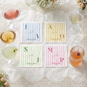 May include: Four square paper napkins with vertical stripes in blue, yellow, green, and pink. Each napkin features two large initials, the word and, and two names with a date printed in the center. The napkins are arranged on a white lace tablecloth.
