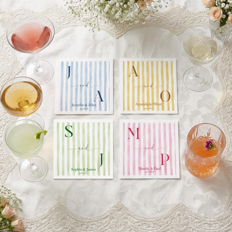 May include: Four square paper napkins with vertical stripes in blue, yellow, green, and pink. Each napkin features two large initials, the word and, and two names with a date printed in the center. The napkins are arranged on a white lace tablecloth.