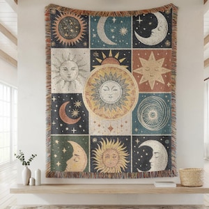 May include: A woven tapestry featuring a celestial design with suns, moons, and stars in a grid pattern. The color palette includes beige, blue, and orange hues. The tapestry has a fringed edge and is displayed on a wall.
