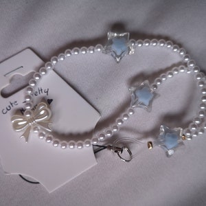 Peut inclure: Un porte-clés de téléphone en perles blanches avec un charme d'étoile bleue et un nœud blanc. Le charme est attaché à une dragonne de téléphone transparente avec un anneau métallique.