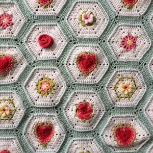 May include: A crocheted blanket with a hexagonal pattern featuring a variety of colorful flowers and hearts. The blanket is made with white, green, pink, red, and yellow yarn.