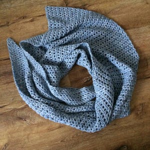 May include: A light blue crocheted scarf with a delicate openwork pattern. The scarf is folded and laid out on a wooden surface.