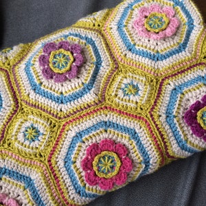 May include: A crocheted blanket with a geometric pattern featuring pink, green, and blue flowers. The blanket is made with a variety of stitches and textures.