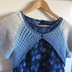 Puede incluir: Una estola de ganchillo azul claro con un diseño degradado de blanco y azul claro. La estola está drapeada sobre un vestido azul con un estampado de árboles blancos.